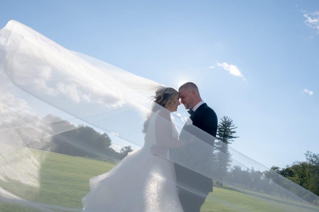 A couple in wedding attire stands closely in an open field with the woman's veil flowing in the breeze under a clear blue sky.